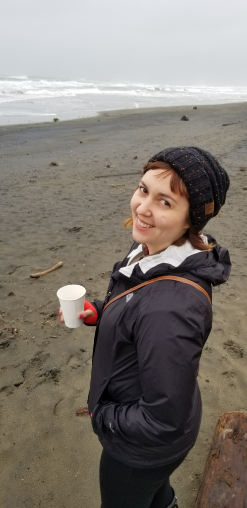 Chelsea Maralason standing on a shoreline in cool weather wearing winter clothing, holding a paper cup